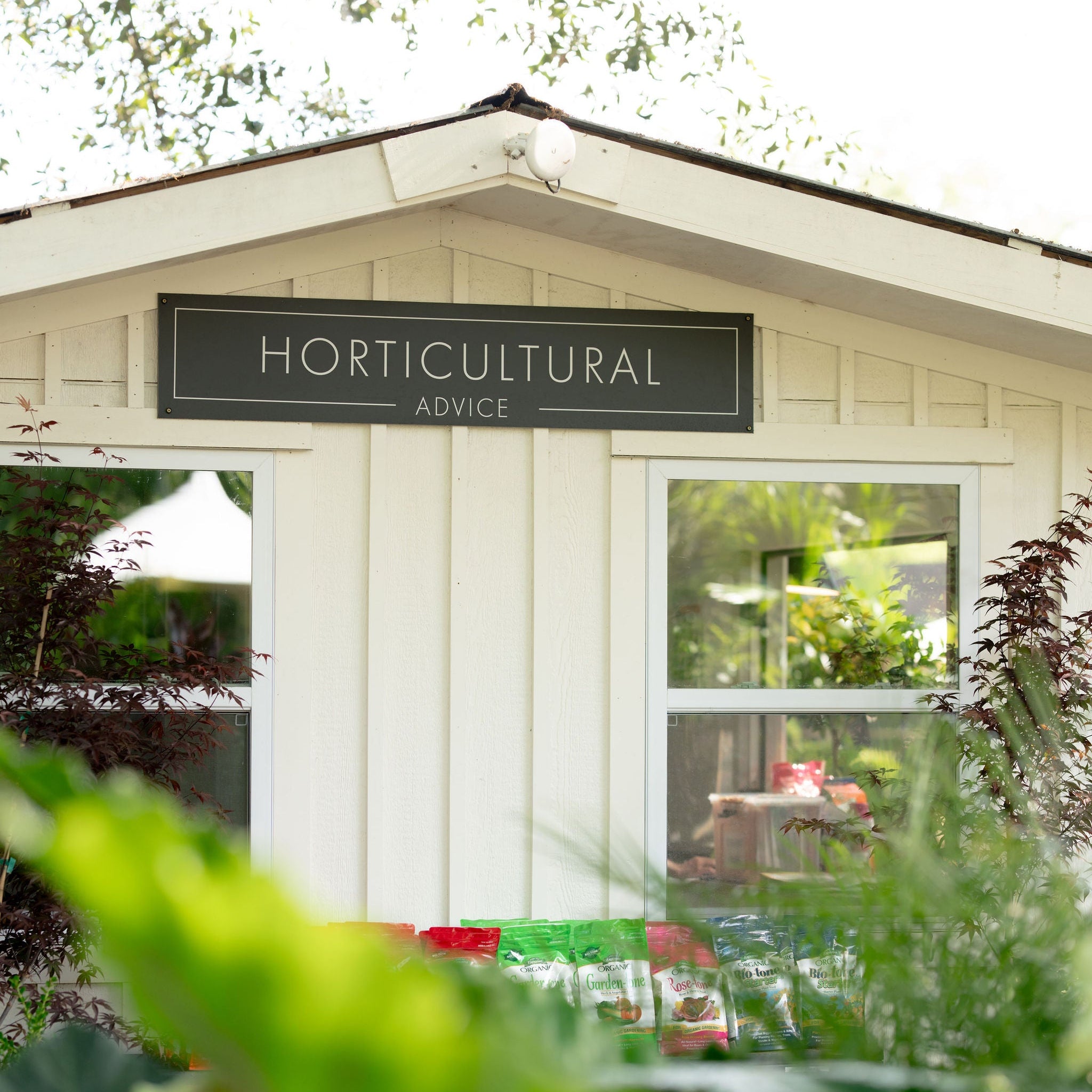 A white wooden garden shed with a black sign reading 'HORTICULTURAL ADVICE' above two windows, surrounded by green garden foliage in the foreground and background.