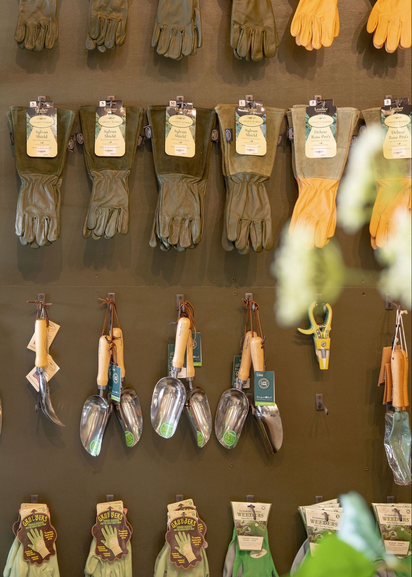 A display of gardening tools and gloves hanging on a rustic wooden wall display inside a garden center. The display features rows of gloves in various colors and sizes, along with hand tools such as trowels, shears, and pruners, under a warm hanging light.