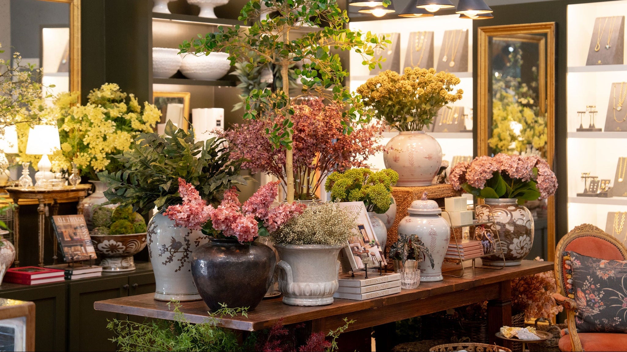 A styled interior scene showing a wooden display table filled with assorted home decor, vases, flowers, books, and decorative objects in a boutique or showroom setting. No person is visible, but the real environment is present, so this fits Lifestyle rather than Product/Packaging.