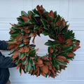 A large magnolia leaf wreath with green and reddish-brown leaves, held against a white garage door by a person whose arm and torso are partially visible on the left.