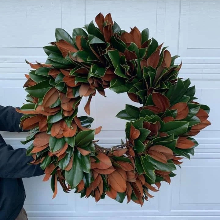 A large magnolia leaf wreath with green and reddish-brown leaves, held against a white garage door by a person whose arm and torso are partially visible on the left.